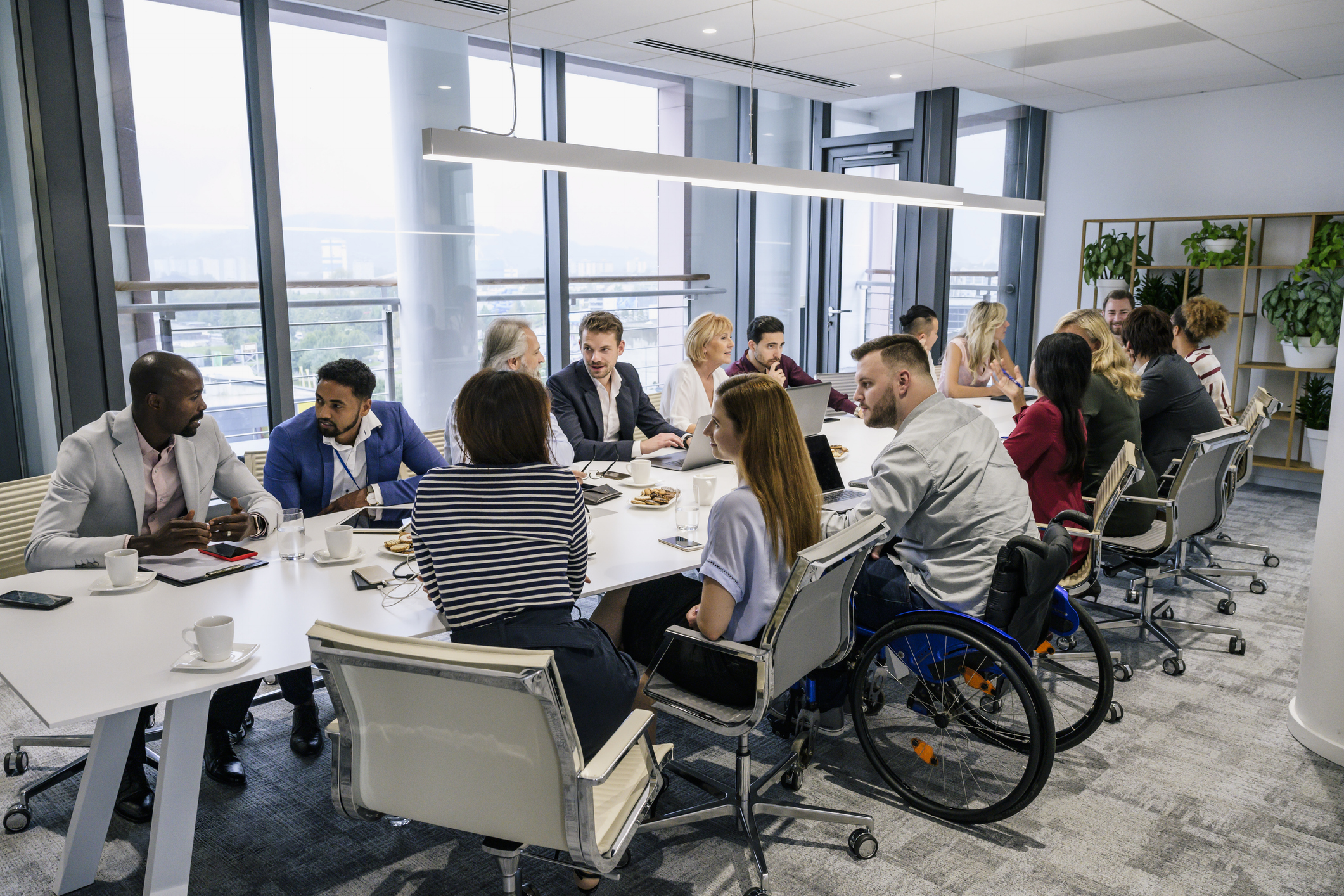 A large meeting room hosting about 15 people of diverse genders and cultures around a table. A person near the front is in a wheelchair.