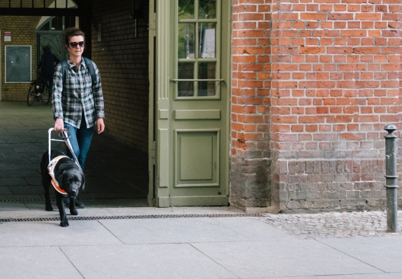 A visually impaired woman walking out of a building with a backpack on and walking with a black lab dog. The dog is guiding her, walking slightly ahead.